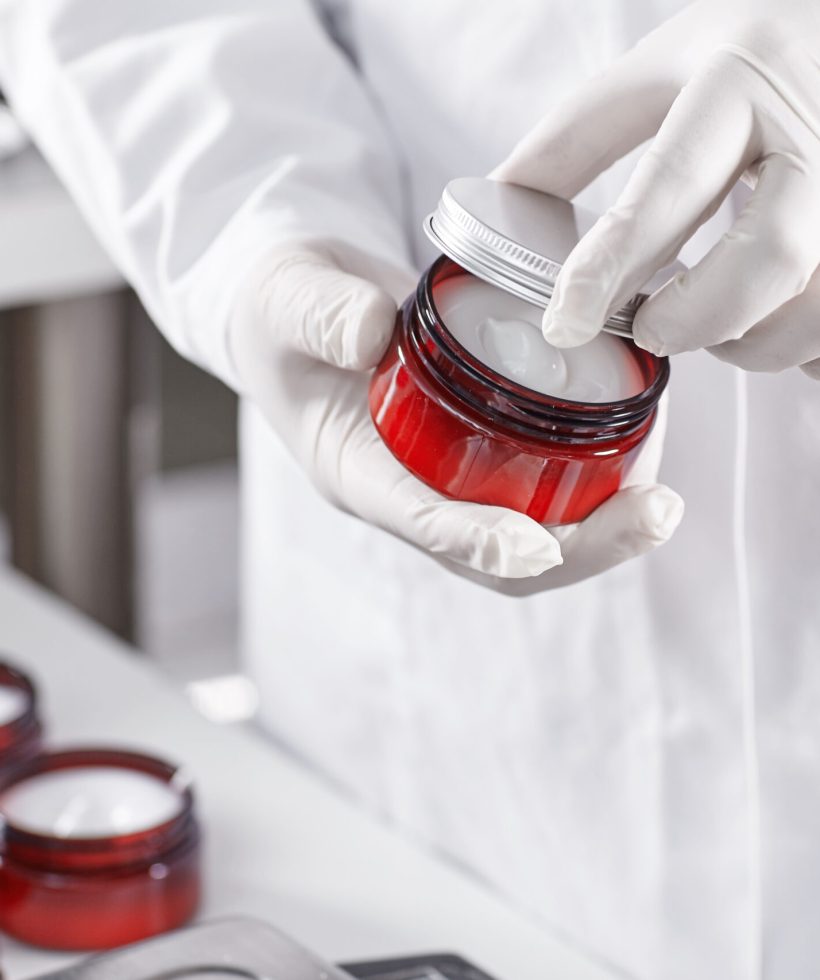 Factory worker in white gown and gloves holding red container with skin care product touching it with finger testing quality. Representing new product on factory. Showing high quality beauty mask